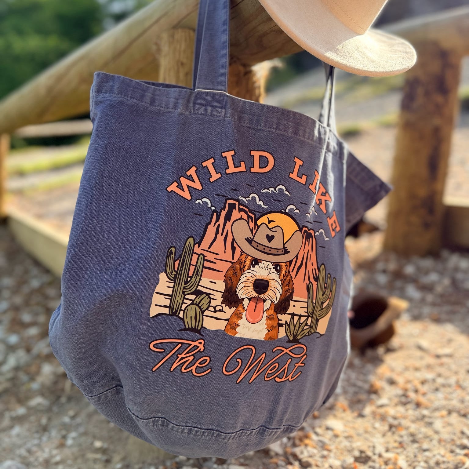 Blue tote bag with 'Wild Like The West' design and cowboy hat on wooden surface
