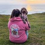 Person in a pink hoodie with a dog, sitting on a grassy hill overlooking the ocean at sunset.