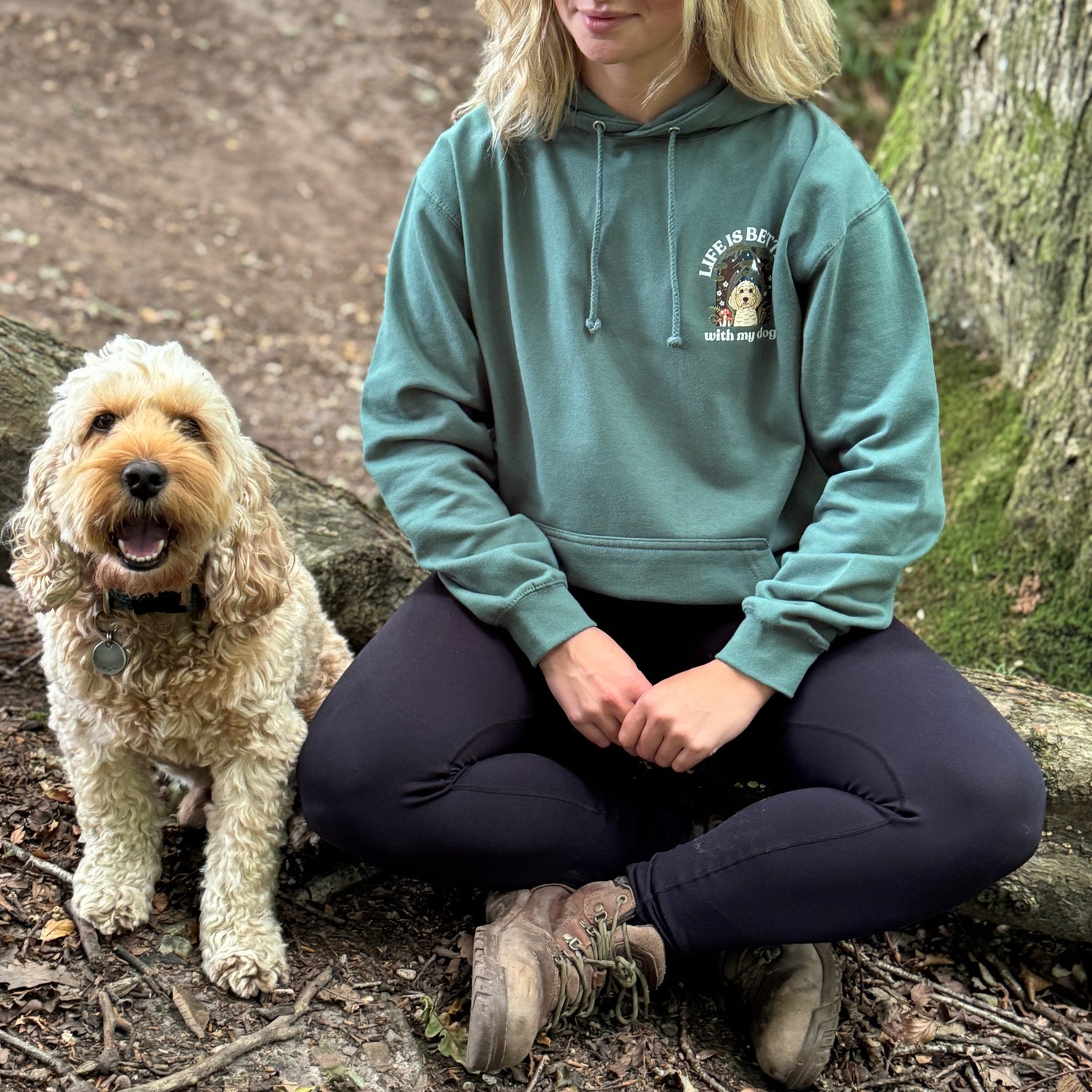 Woman sitting on a forest path with a dog, wearing a green hoodie with a logo.