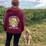 A woman viewed from behind wearing a maroon hoodie with a large graphic on the back that reads 'LIFE IS BETTER with my dog' around a dog and sunflower illustration. She is standing in a sunflower field next to a fluffy light brown dog under a blue sky with clouds.