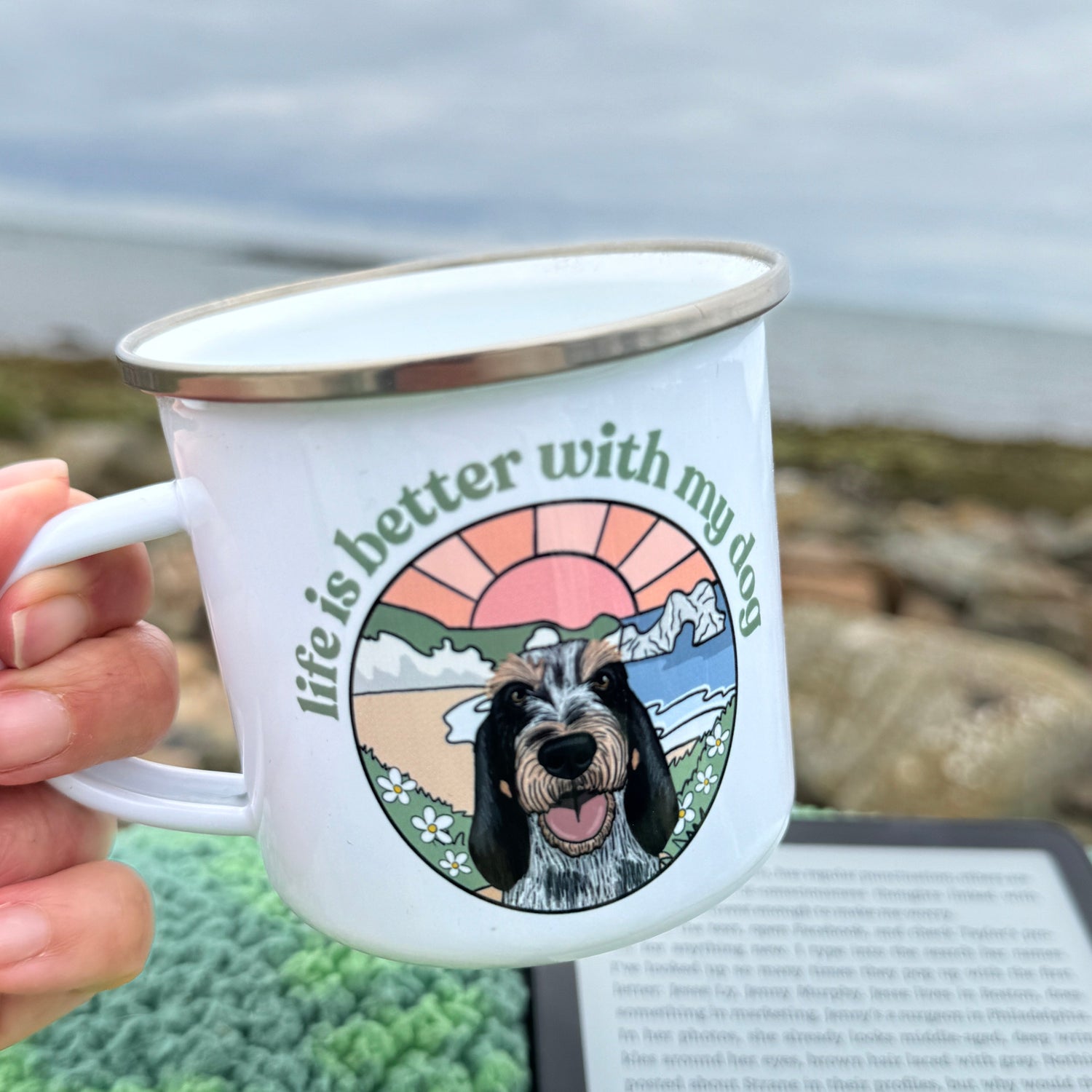 White mug with a dog illustration and text held by a hand, against a blurred background.