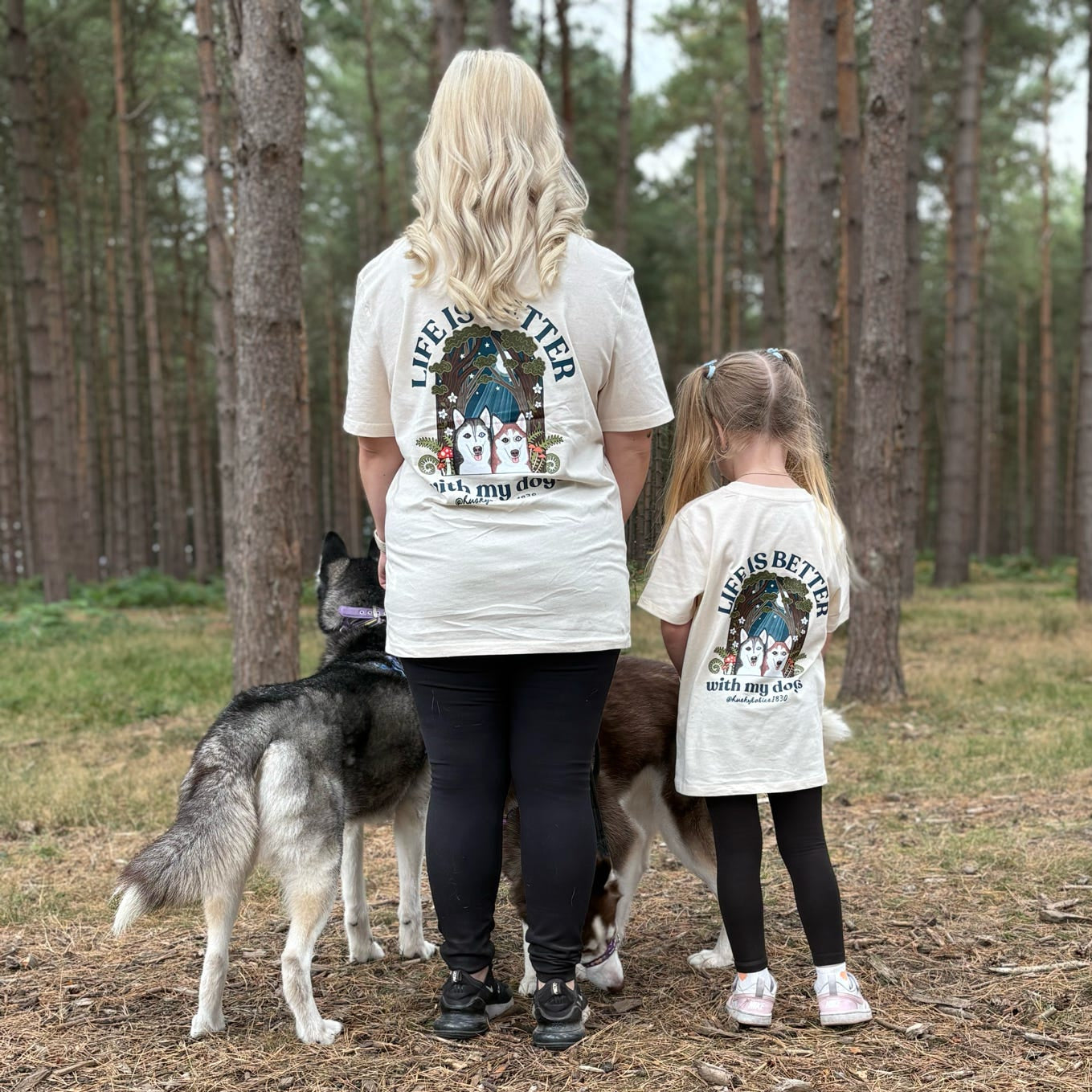 Two people and two dogs standing in a forest setting wearing matching t-shirts.