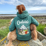 Person wearing a teal hoodie with 'Outside is Where it's At' text and nature-themed design, sitting on rocks by the sea.