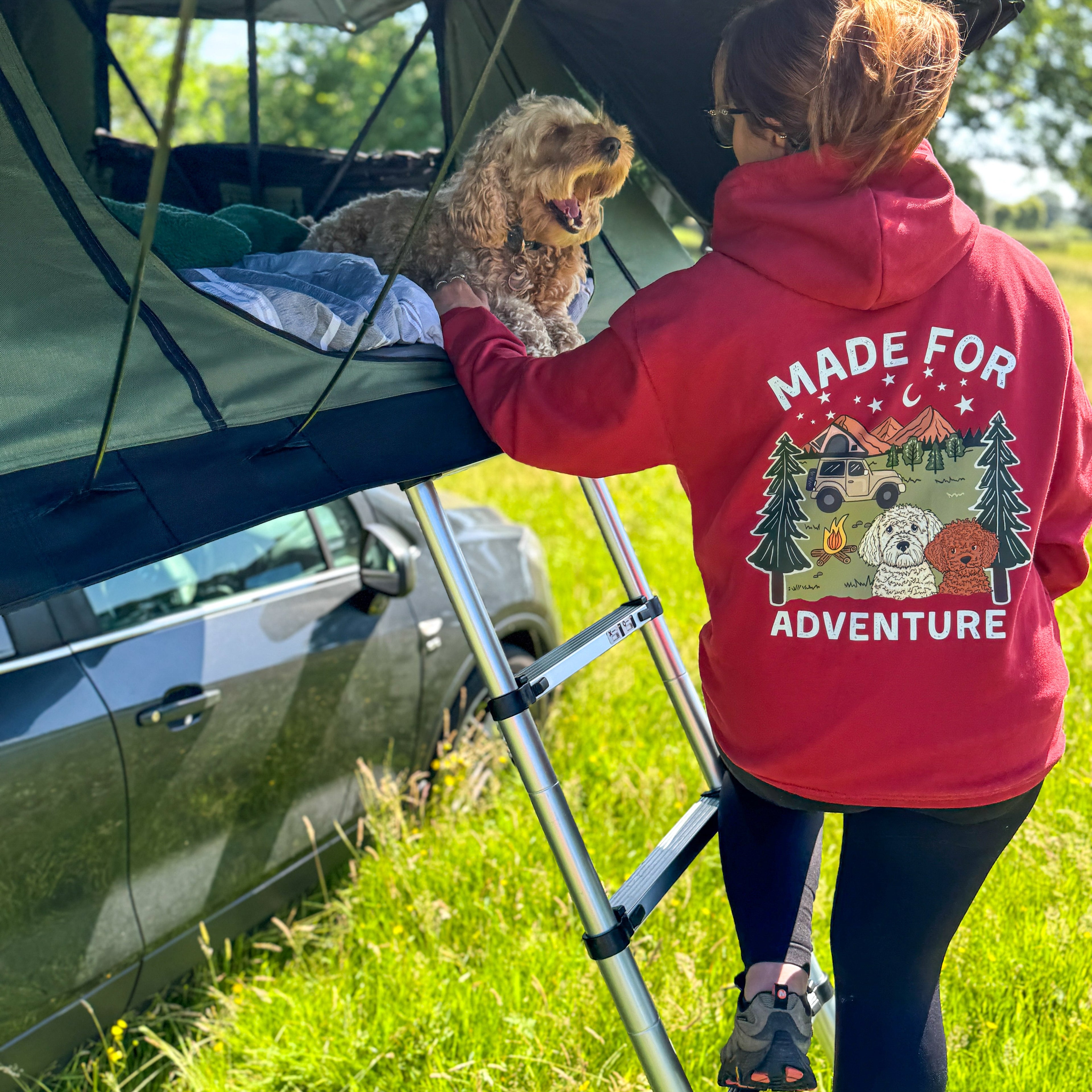 Person in a red 'Made for Adventure' hoodie with a dog on a vehicle roof tent in a grassy area.