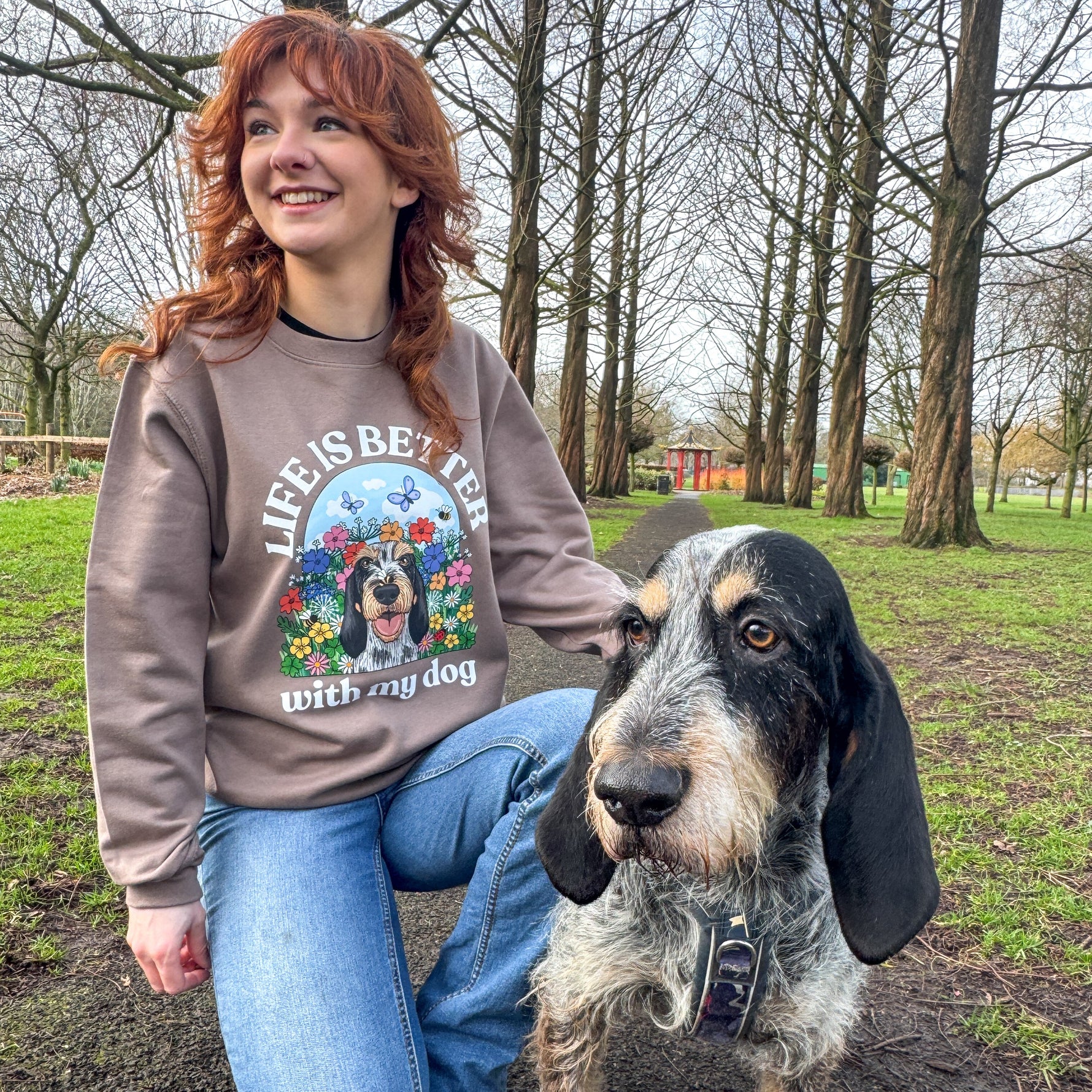 Person wearing a sweatshirt with a dog graphic and text, kneeling next to a dog in an outdoor setting.