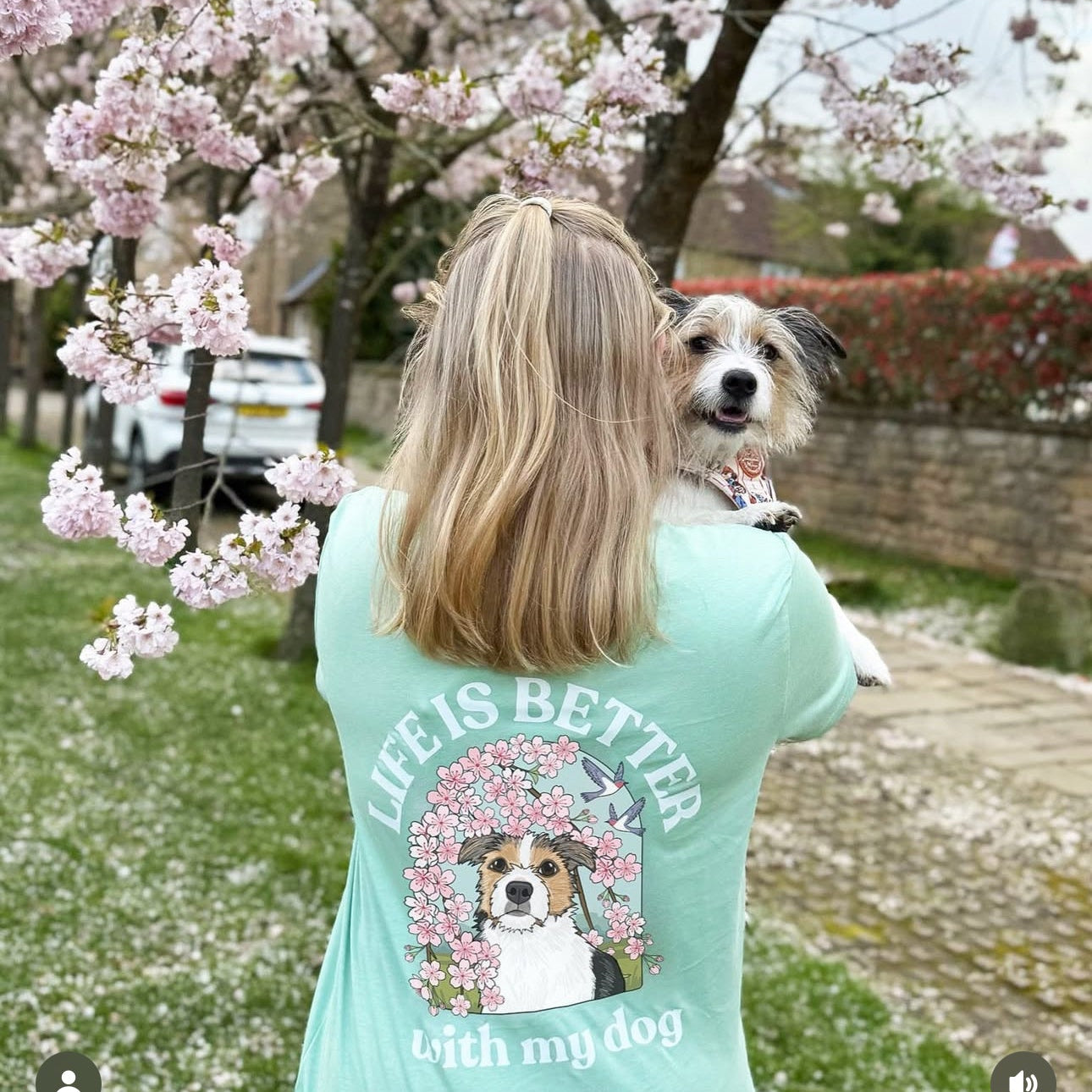 Woman wearing a teal t-shirt with a dog graphic and 'Life is better with my dog' text, standing in a park with cherry blossoms.