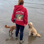 Person wearing a red hoodie with 'Made for Adventure' text on a beach with two dogs.