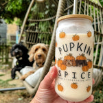 A hand holding a frosted glass tumbler with 'PUPKIN SPICE' text and pumpkin illustrations, accompanied by a reusable metal straw and a wooden bamboo lid, with two dogs sitting in a hammock in the background.