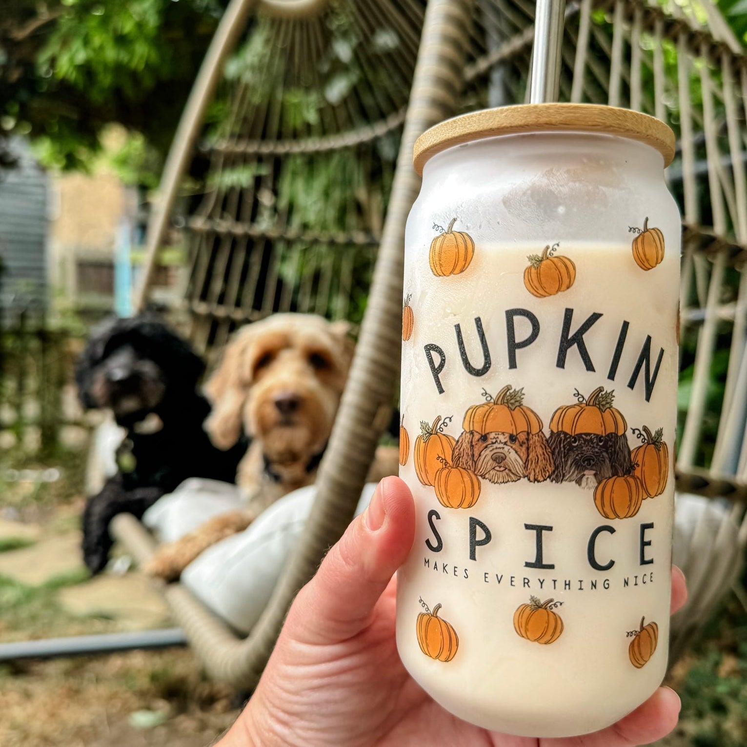 A hand holding a frosted glass tumbler with 'PUPKIN SPICE' text and pumpkin illustrations, accompanied by a reusable metal straw and a wooden bamboo lid, with two dogs sitting in a hammock in the background.