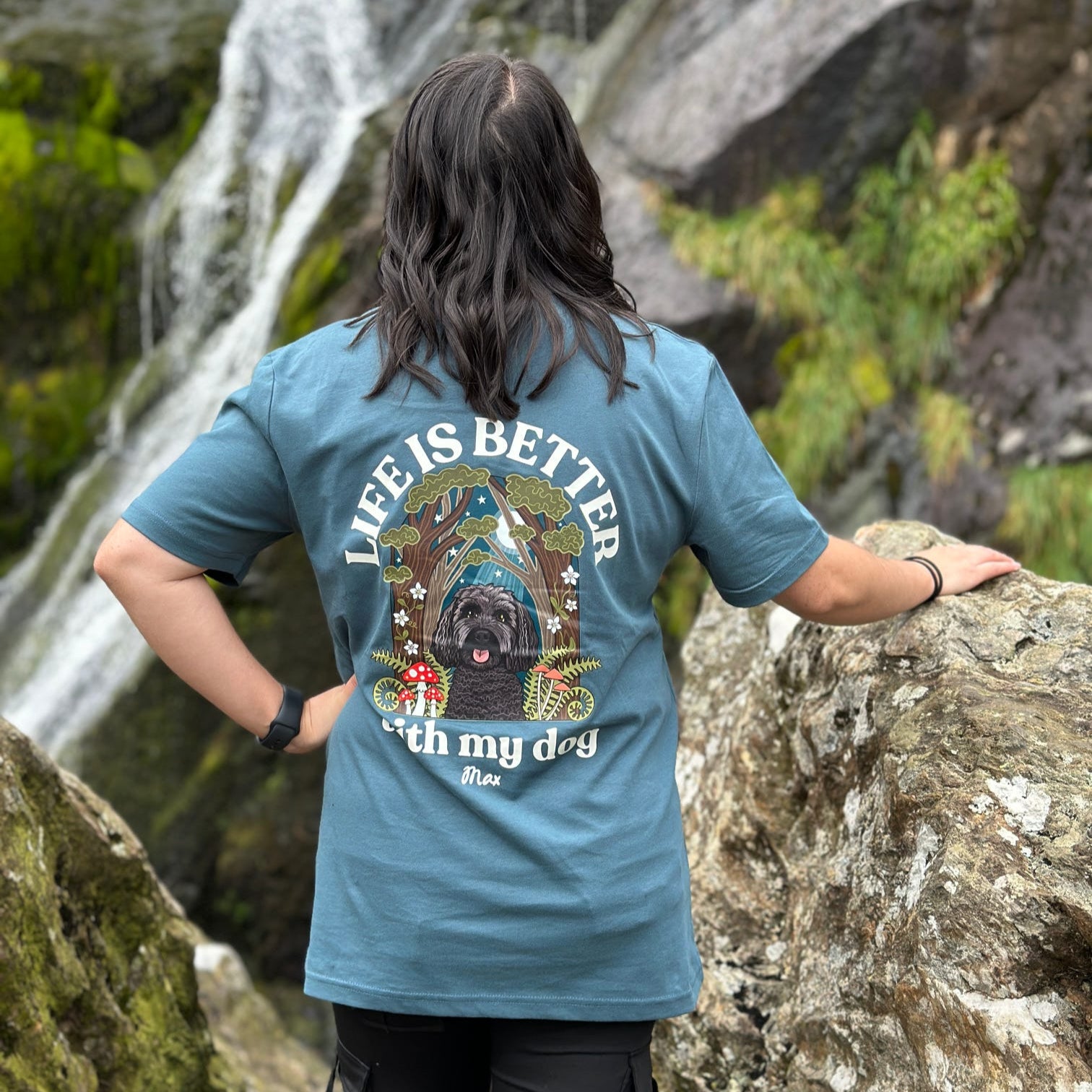 Person wearing a blue t-shirt with a dog graphic and 'Life is better with my dog' text, standing in front of a waterfall.