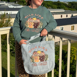 Person wearing a sweatshirt and holding a tote bag with 'Wild Like The West' design on a balcony.