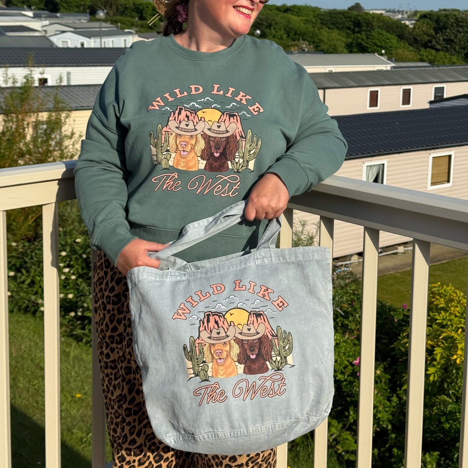 Person wearing a sweatshirt and holding a tote bag with 'Wild Like The West' design on a balcony.